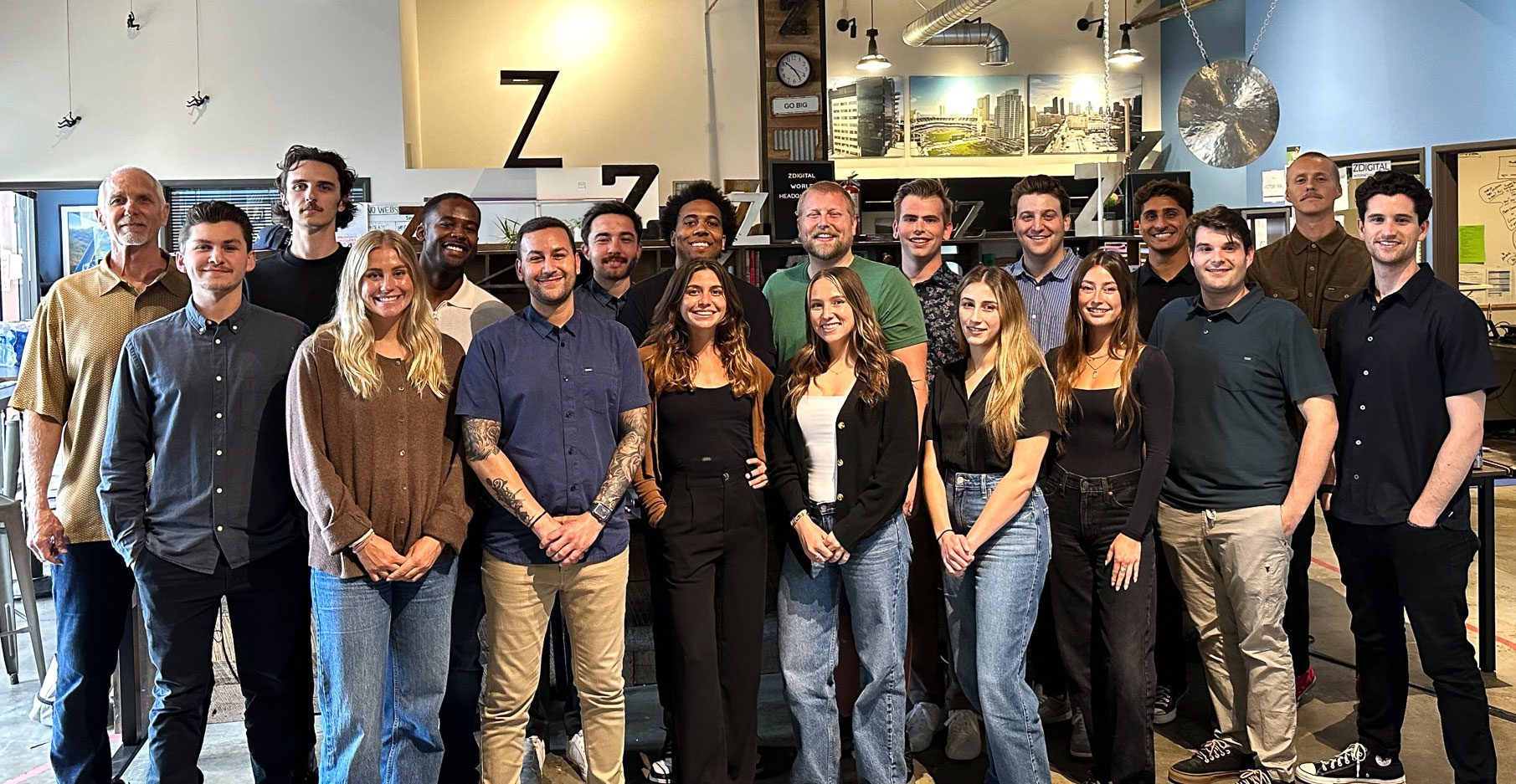 A group of 20 people poses for a photo in an office setting, with a mix of men and women standing and smiling at the camera.