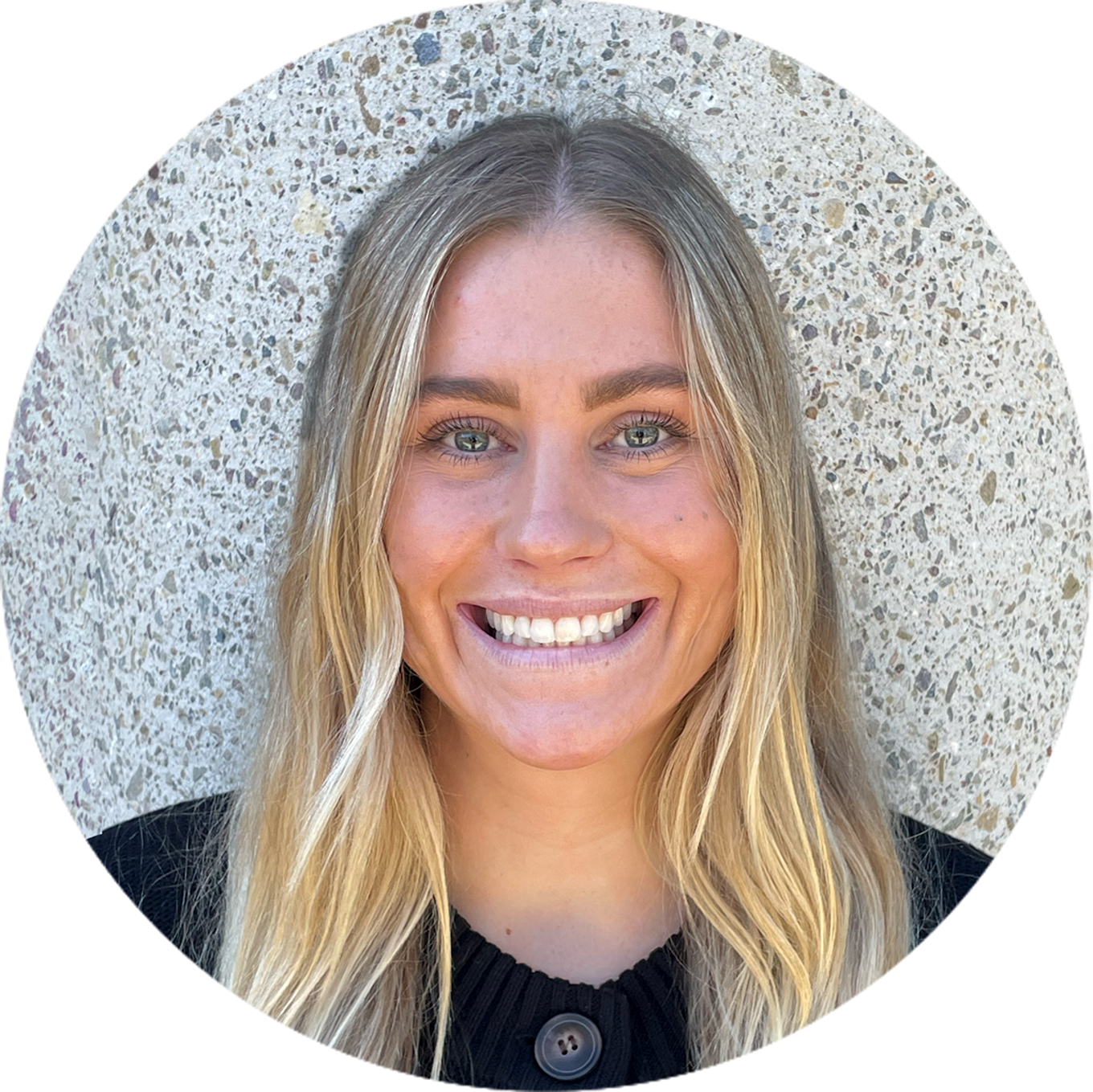 Smiling person with long blonde hair and a black shirt stands in front of a textured concrete wall.